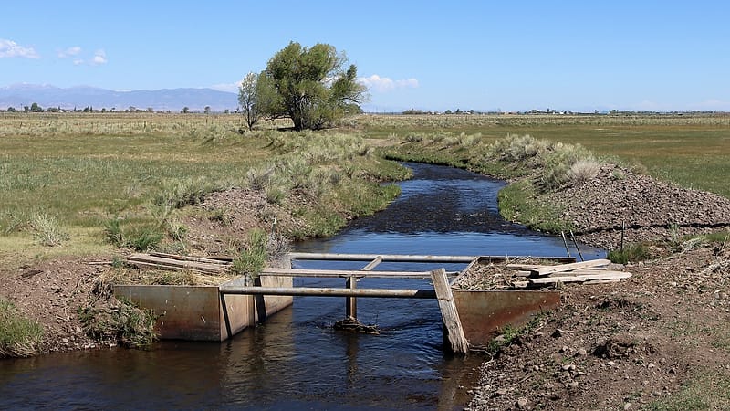 A collapsed wooden flume and a silted ditch overgrown with dry grass running across a fenced field toward hills.