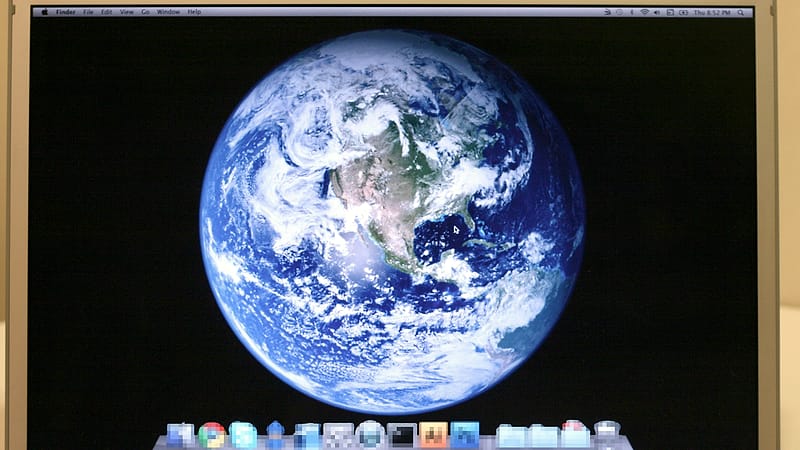 A hand holding a smartphone showing a plain white screen of small text, with a laptop out of focus behind it.