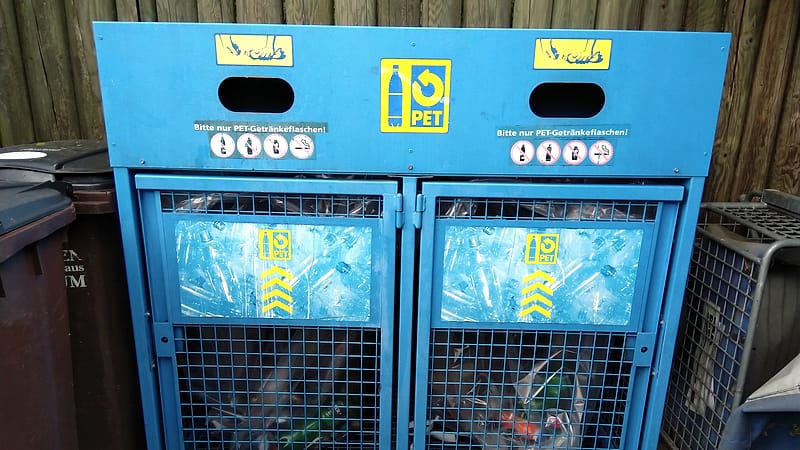 A large outdoor collection container for used plastic drink bottles standing on a paved surface beside a low wall.