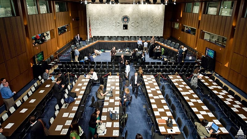 A hearing room with a plain table, a water pitcher, three chairs and a stenographer's machine on a stand.
