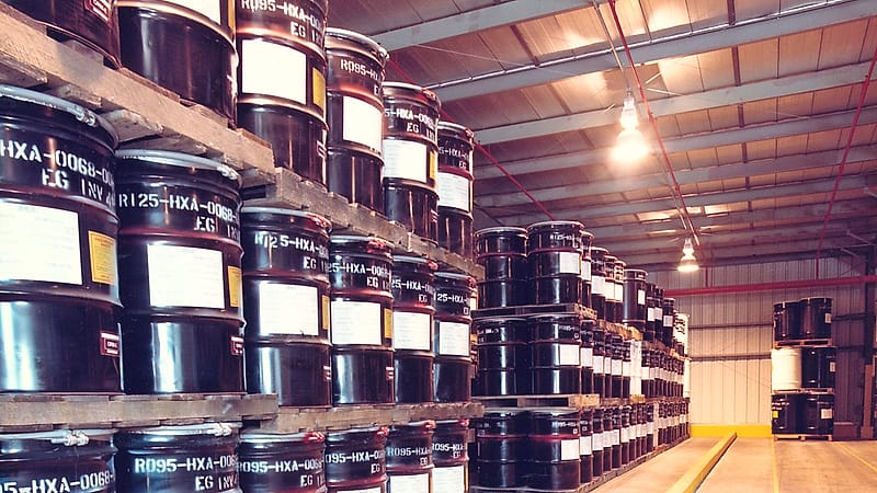 Steel drums with diamond warning labels strapped to a pallet inside a warehouse under bright ceiling lights.