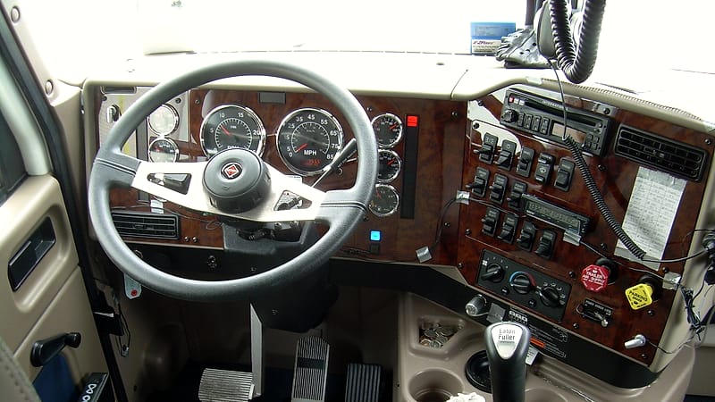 A truck cab interior at dusk showing the steering wheel, gauges and a dark highway through the windshield.