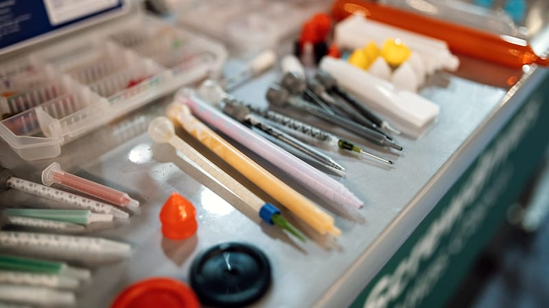 Two people at a workbench in a bright laboratory, one pointing at equipment while the other takes notes.