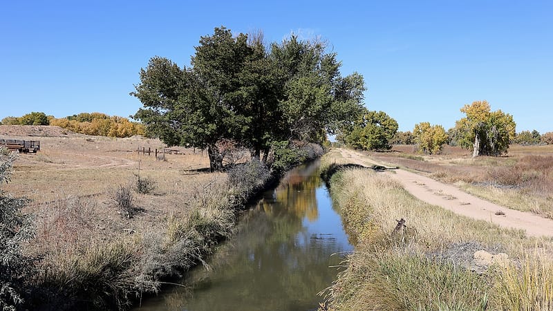 A concrete headgate on an earthen ditch with water running through it beside a dry field and low hills behind.