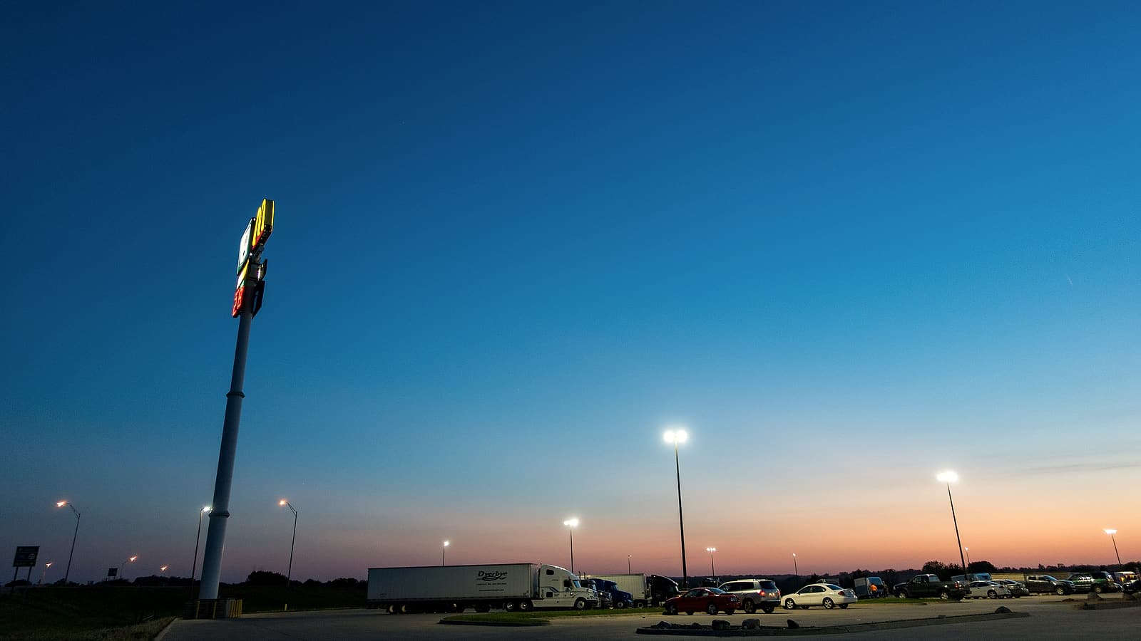 A sleeper tractor with a personalized cab parked at a truck stop beside a fuel island at first light.