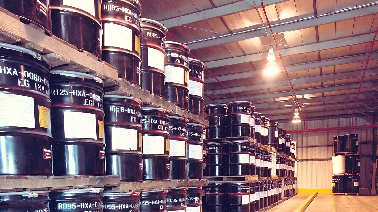Steel drums with diamond warning labels strapped to a pallet inside a warehouse under bright ceiling lights.