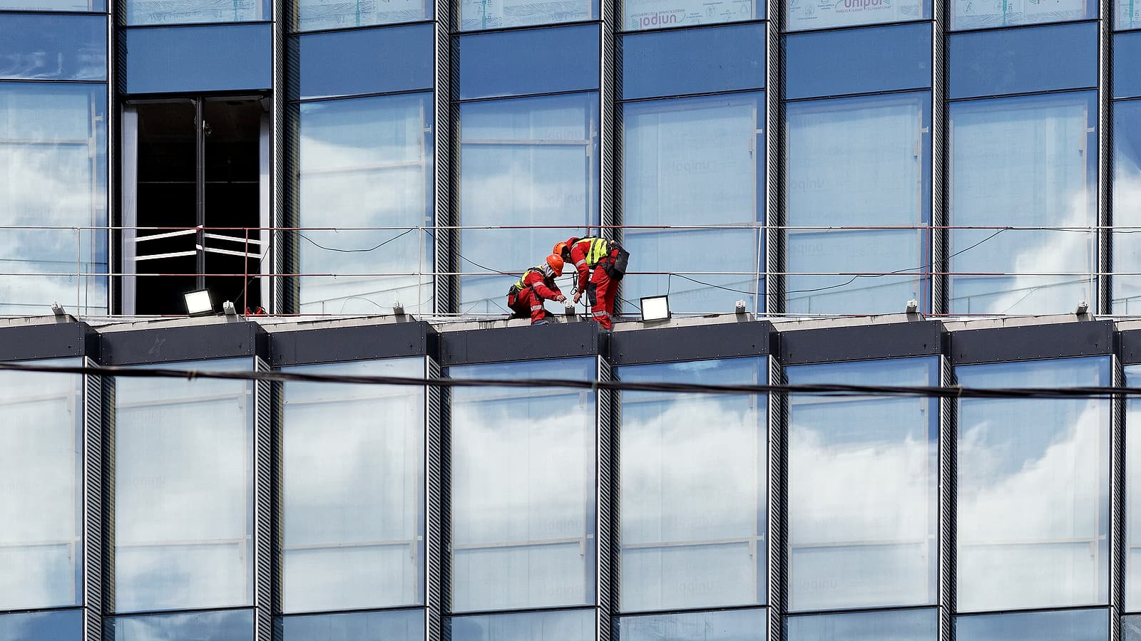 A partly built steel frame on a construction site with two crews working on separate levels under a pale sky.